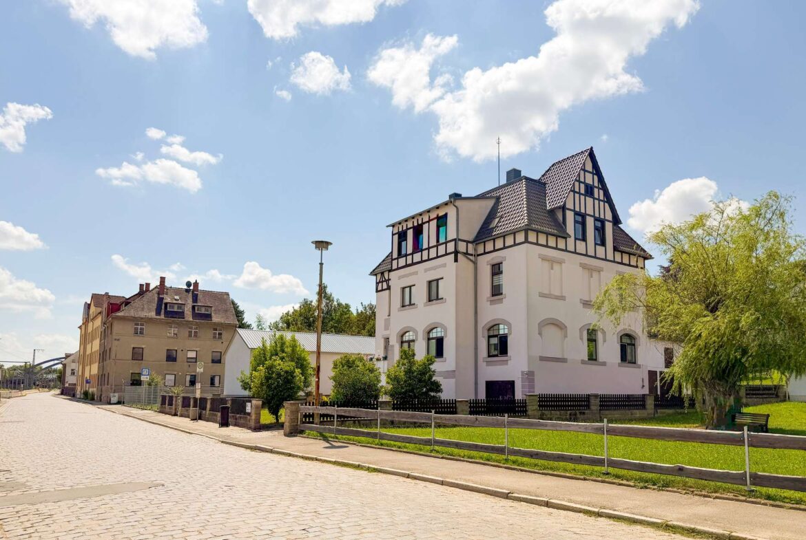 White multi-story building with a steep, timber-framed roof beside a cobblestone street on a sunny day.