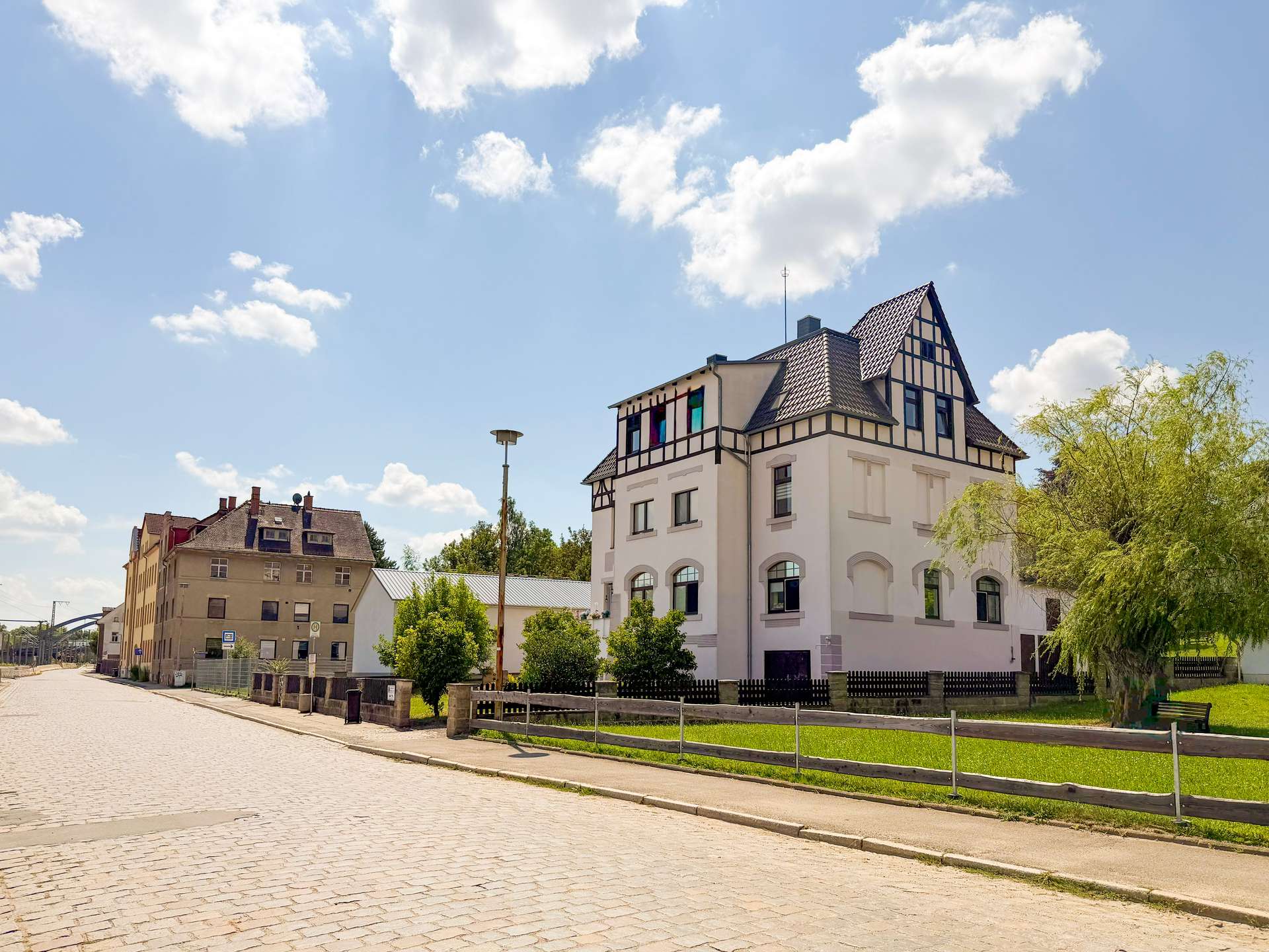 White multi-story building with a steep, timber-framed roof beside a cobblestone street on a sunny day.
