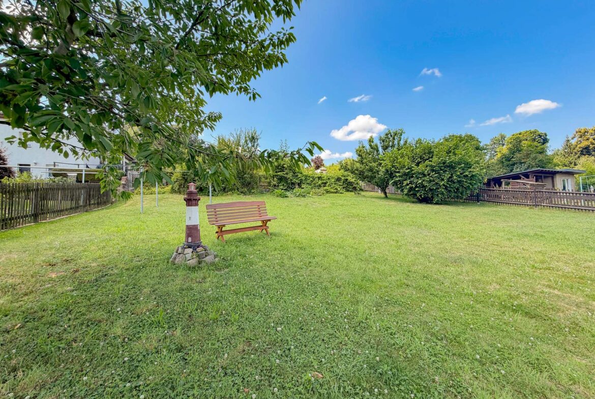 Park scene with a wooden bench beside a small pillar under a leafy overhang, open green lawn and blue sky.