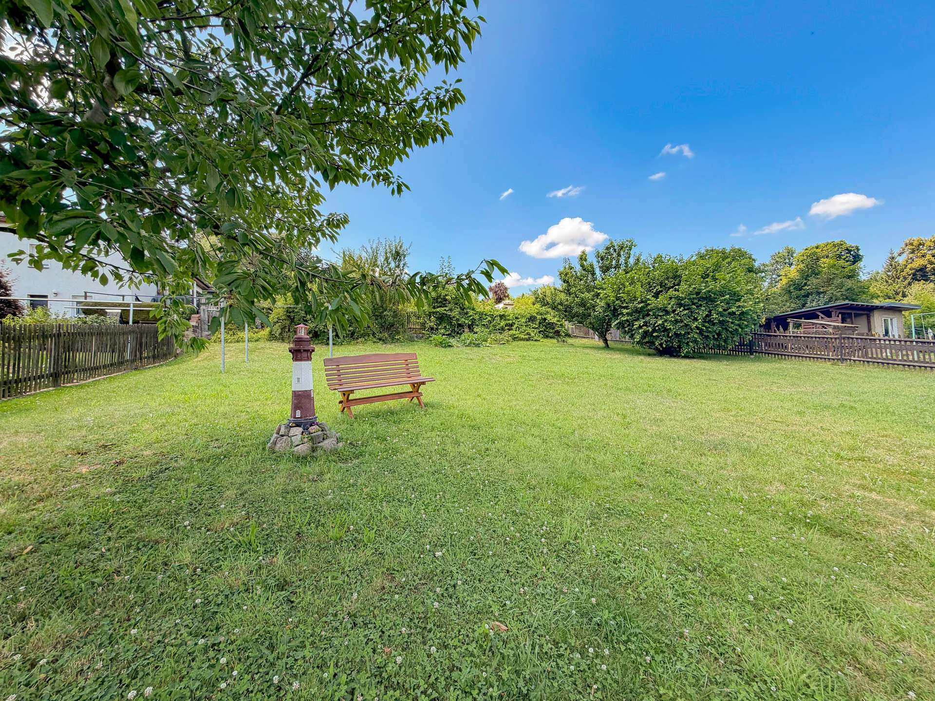 Park scene with a wooden bench beside a small pillar under a leafy overhang, open green lawn and blue sky.