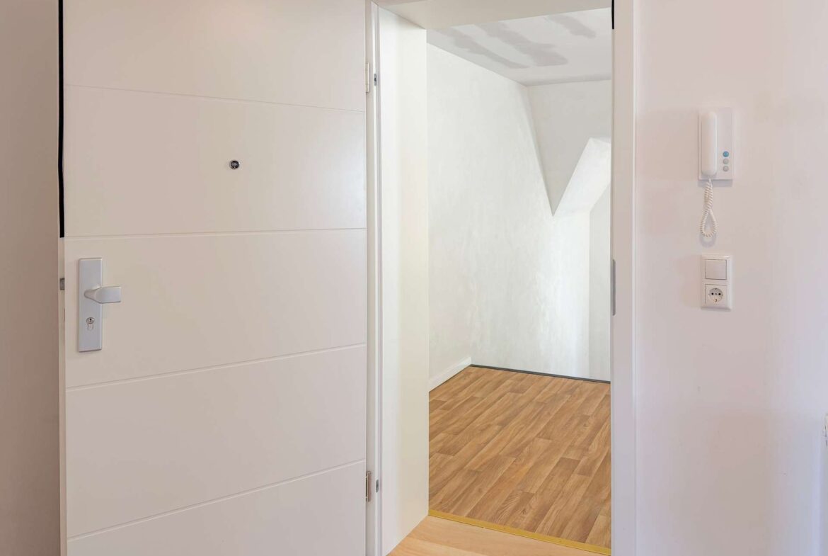 Open white front door revealing a hallway with a wooden floor and a wall-mounted intercom.