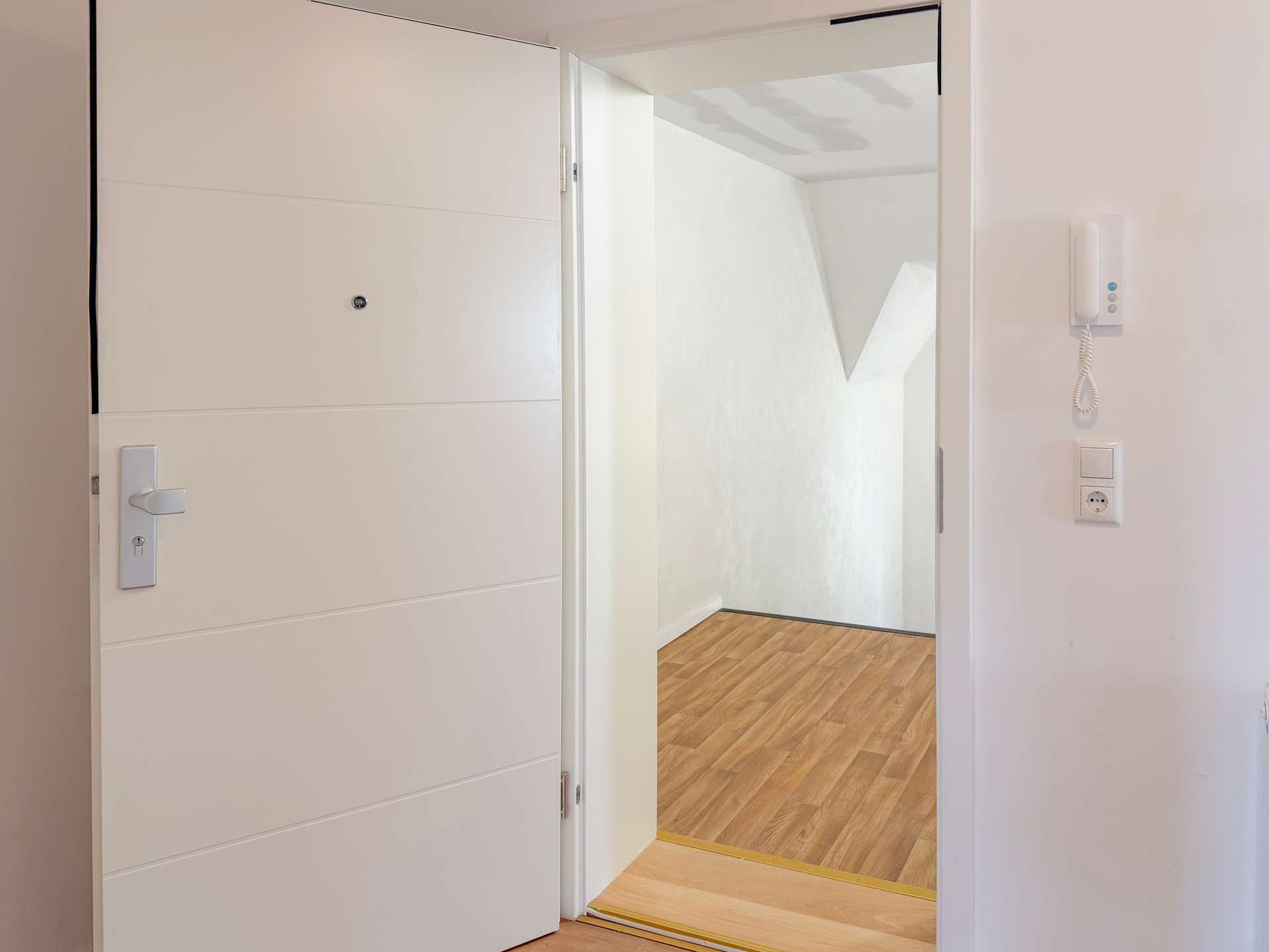 Open white front door revealing a hallway with a wooden floor and a wall-mounted intercom.