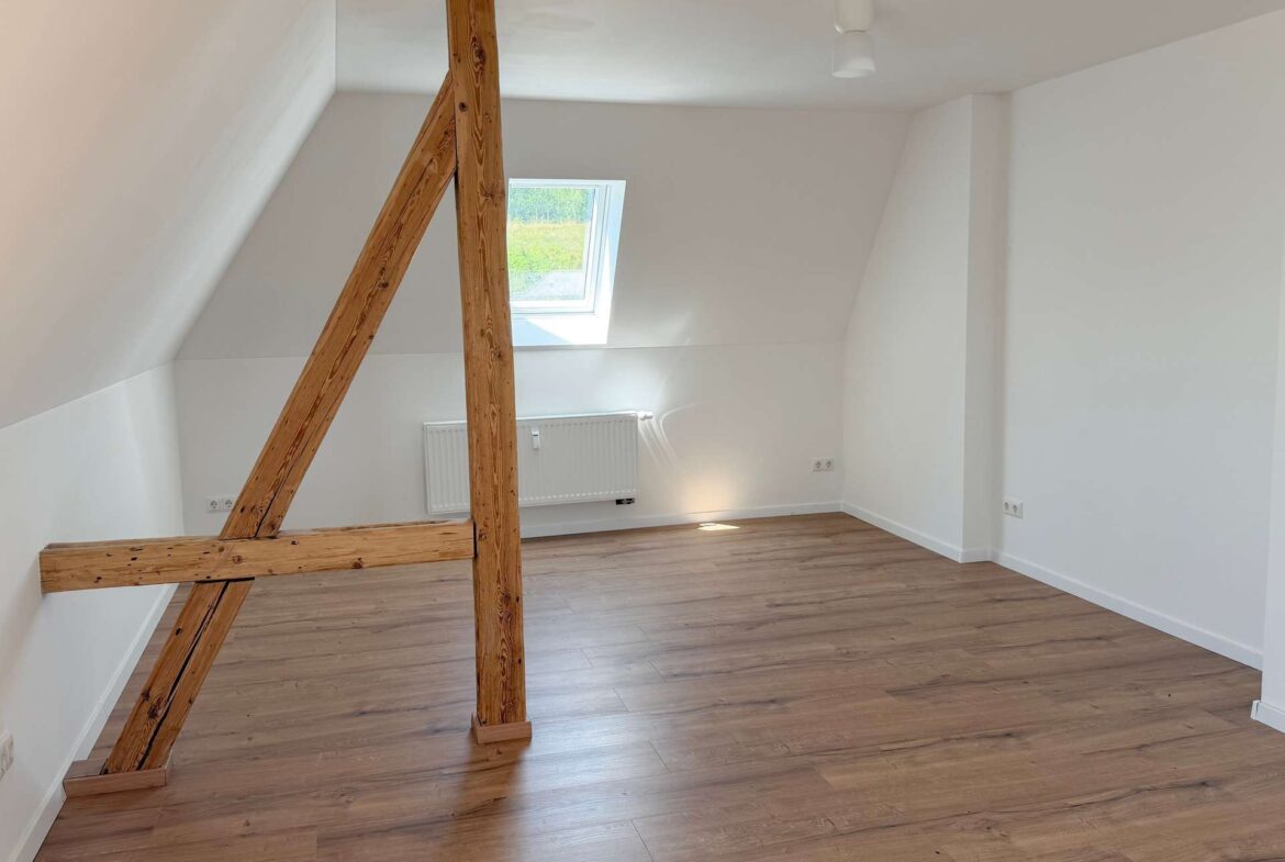 Bright attic room with sloped white walls, exposed wooden beams, a skylight window, and a wood floor.