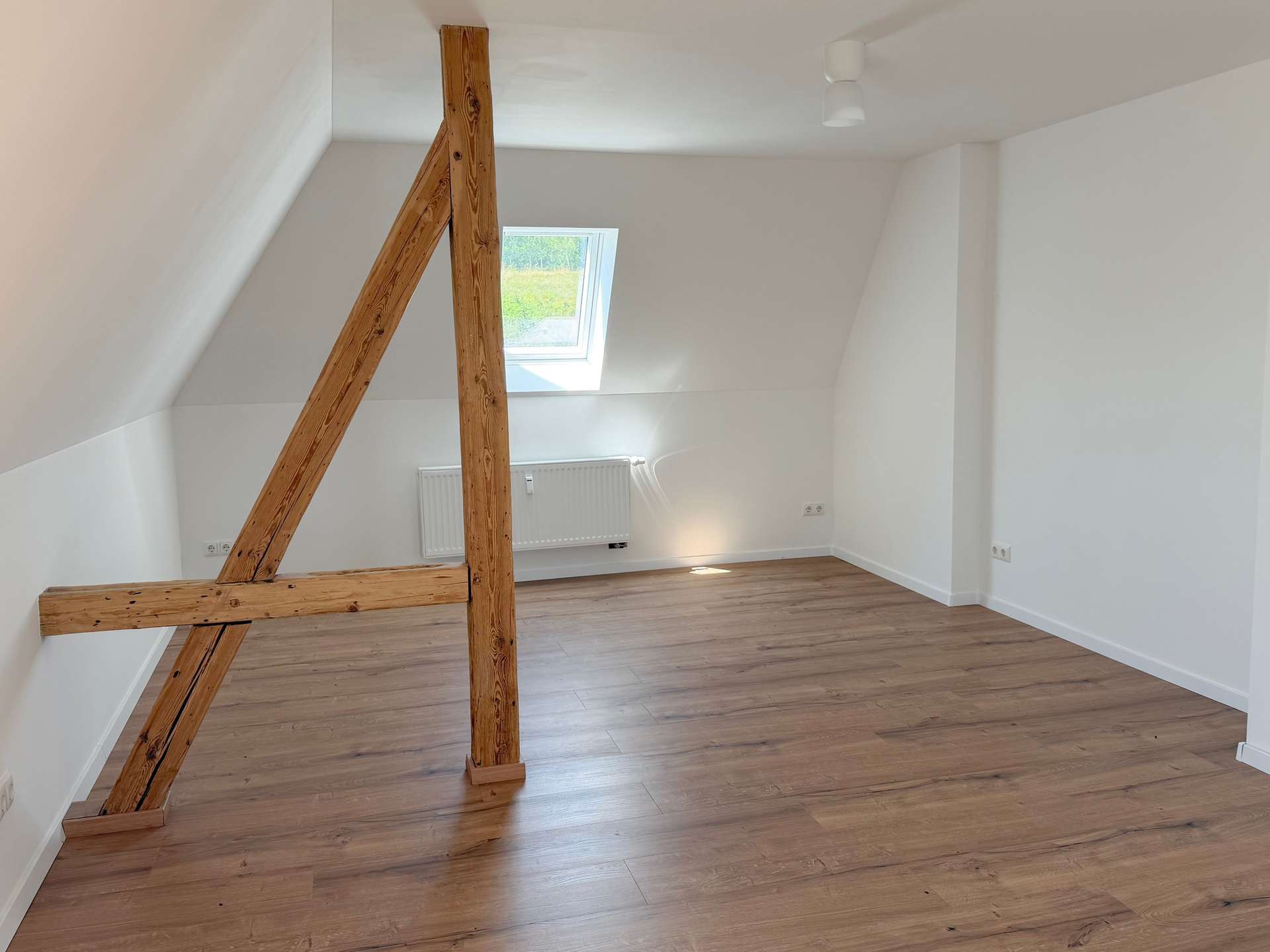 Bright attic room with sloped white walls, exposed wooden beams, a skylight window, and a wood floor.