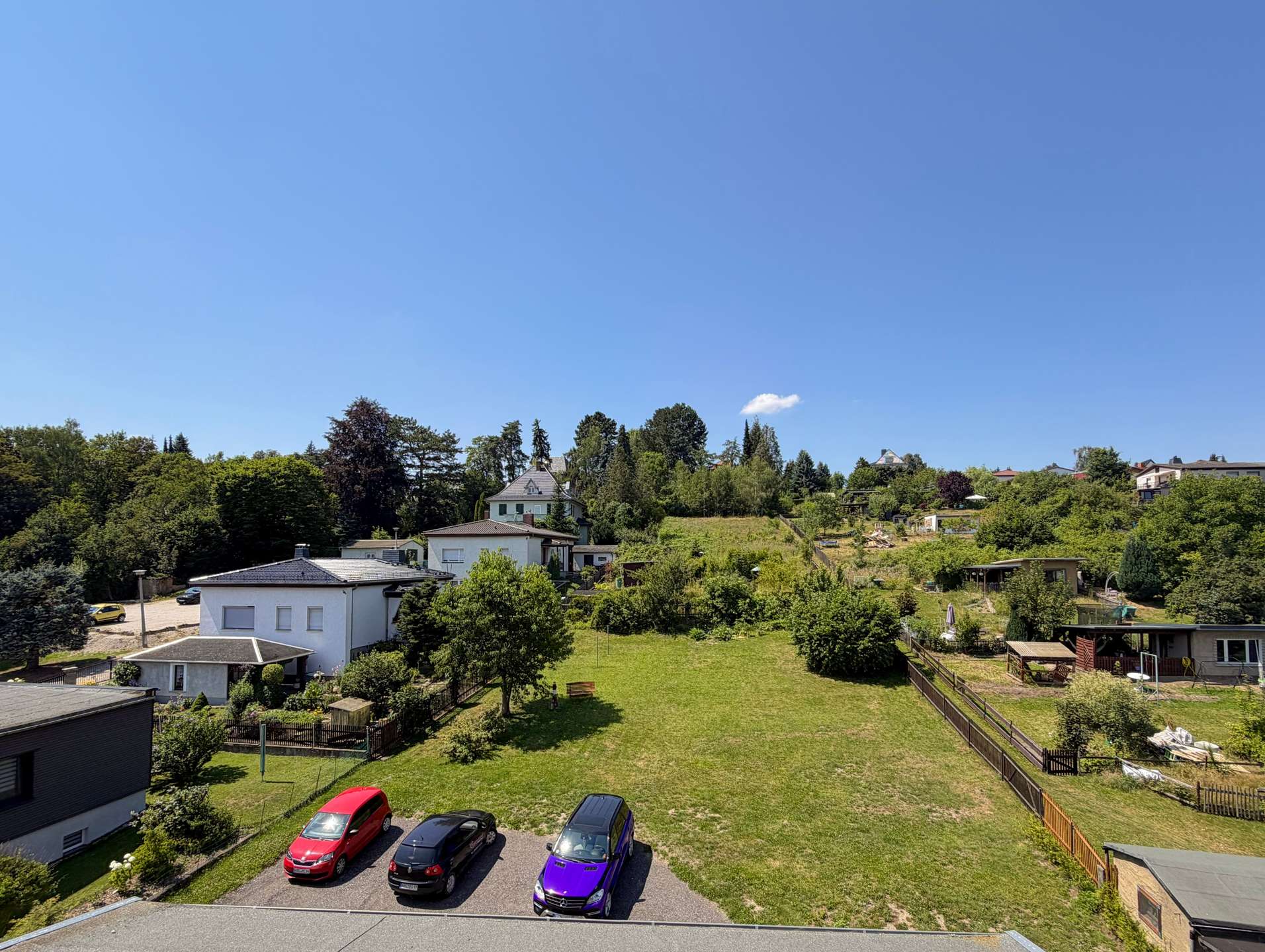 Sunny suburban scene with a row of houses and green lawns under a clear blue sky; several colorful cars parked in a driveway in the foreground.