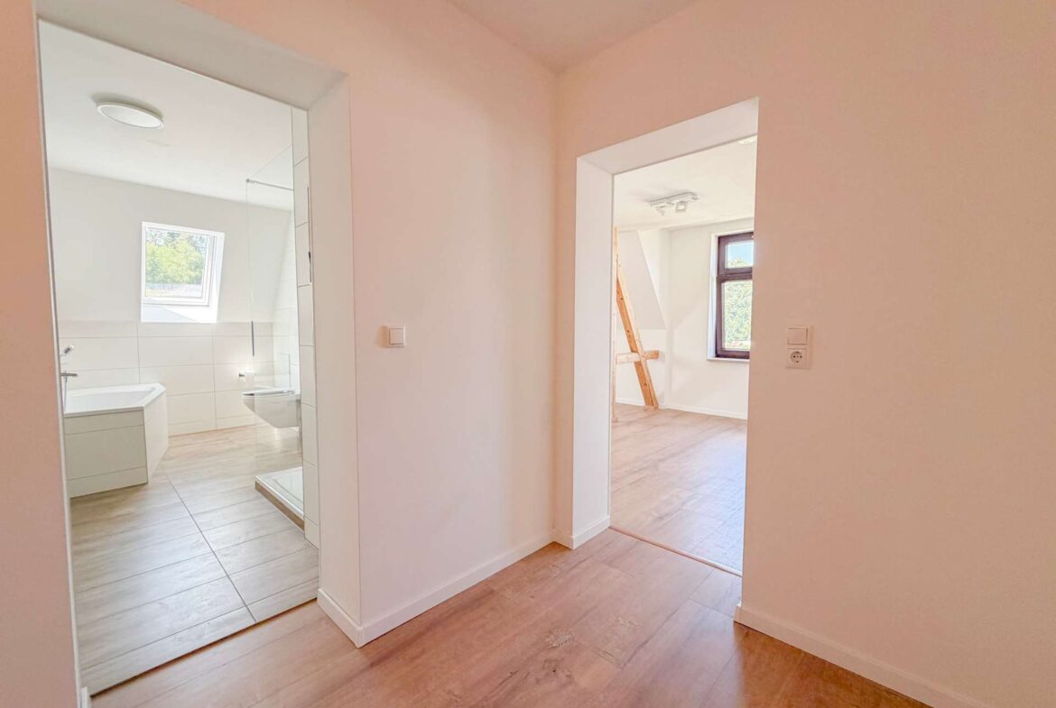 Bright hallway leading to a bathroom with a mirrored cabinet and sink on the left, and a sunlit room with a wooden ladder visible through an open doorway on the right side of the image.