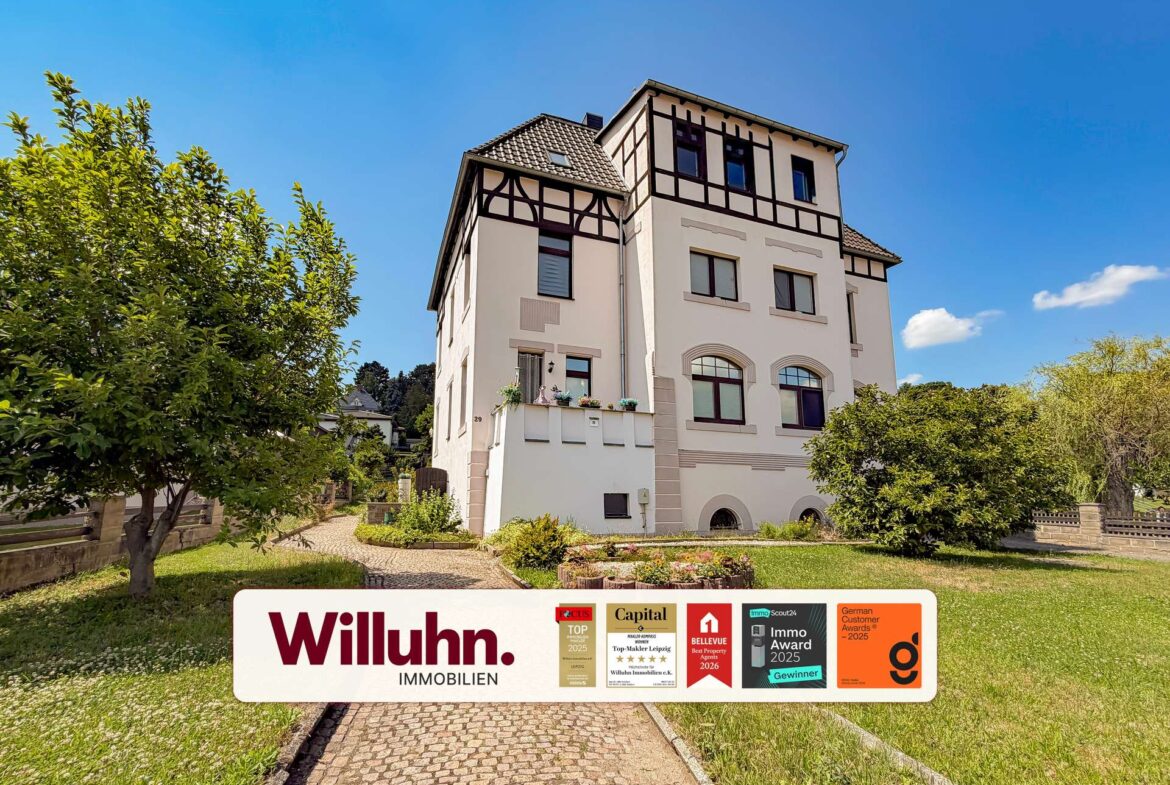 Cream-colored multi-story house with half-timbered detailing, blue sky, and a front garden; a real estate sign reading 'Willuhn Immobilien' dominates the foreground.