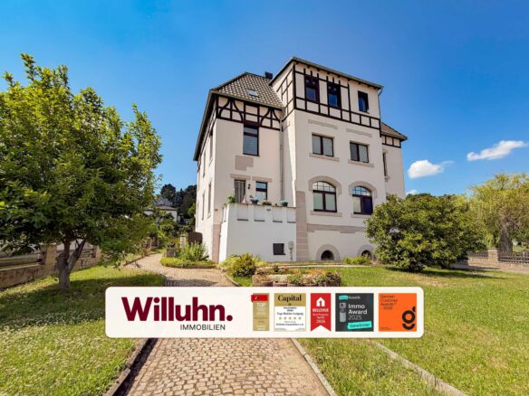 Cream-colored multi-story house with half-timbered detailing, blue sky, and a front garden; a real estate sign reading 'Willuhn Immobilien' dominates the foreground.