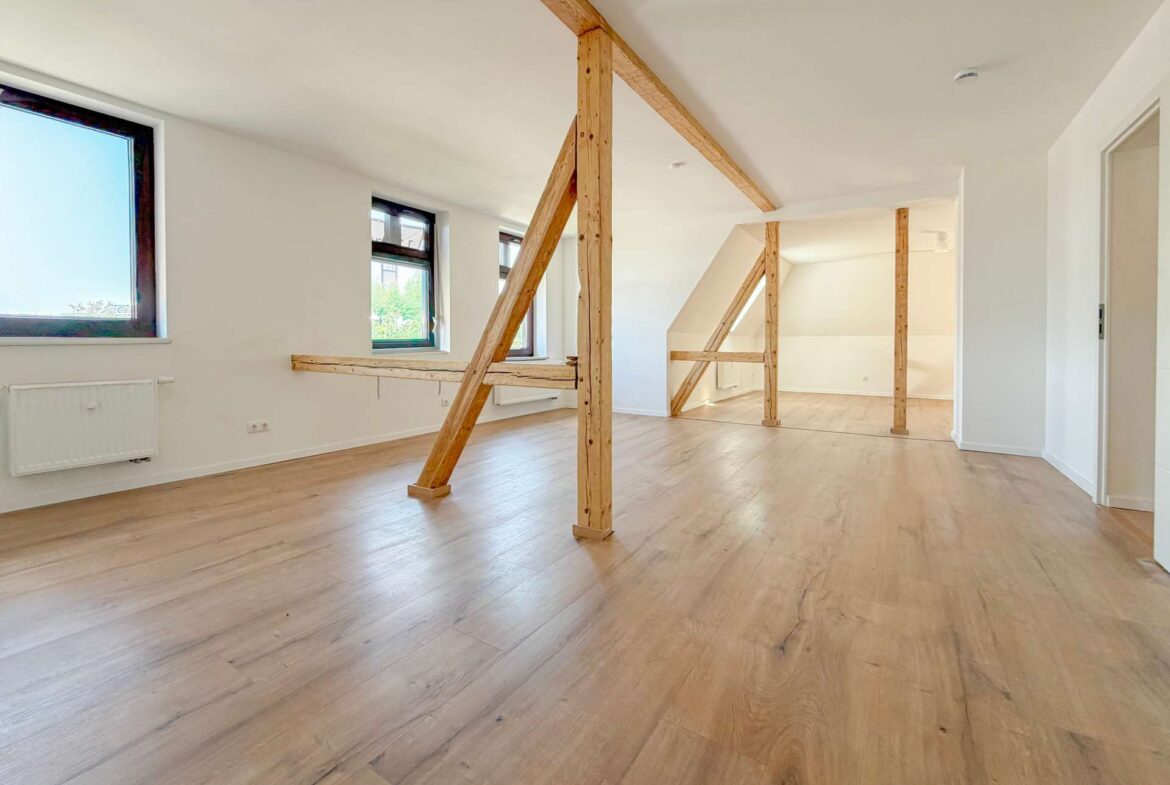 Bright empty loft with light wood floors, white walls, and exposed wooden structural beams running across the ceiling and down the room edge.