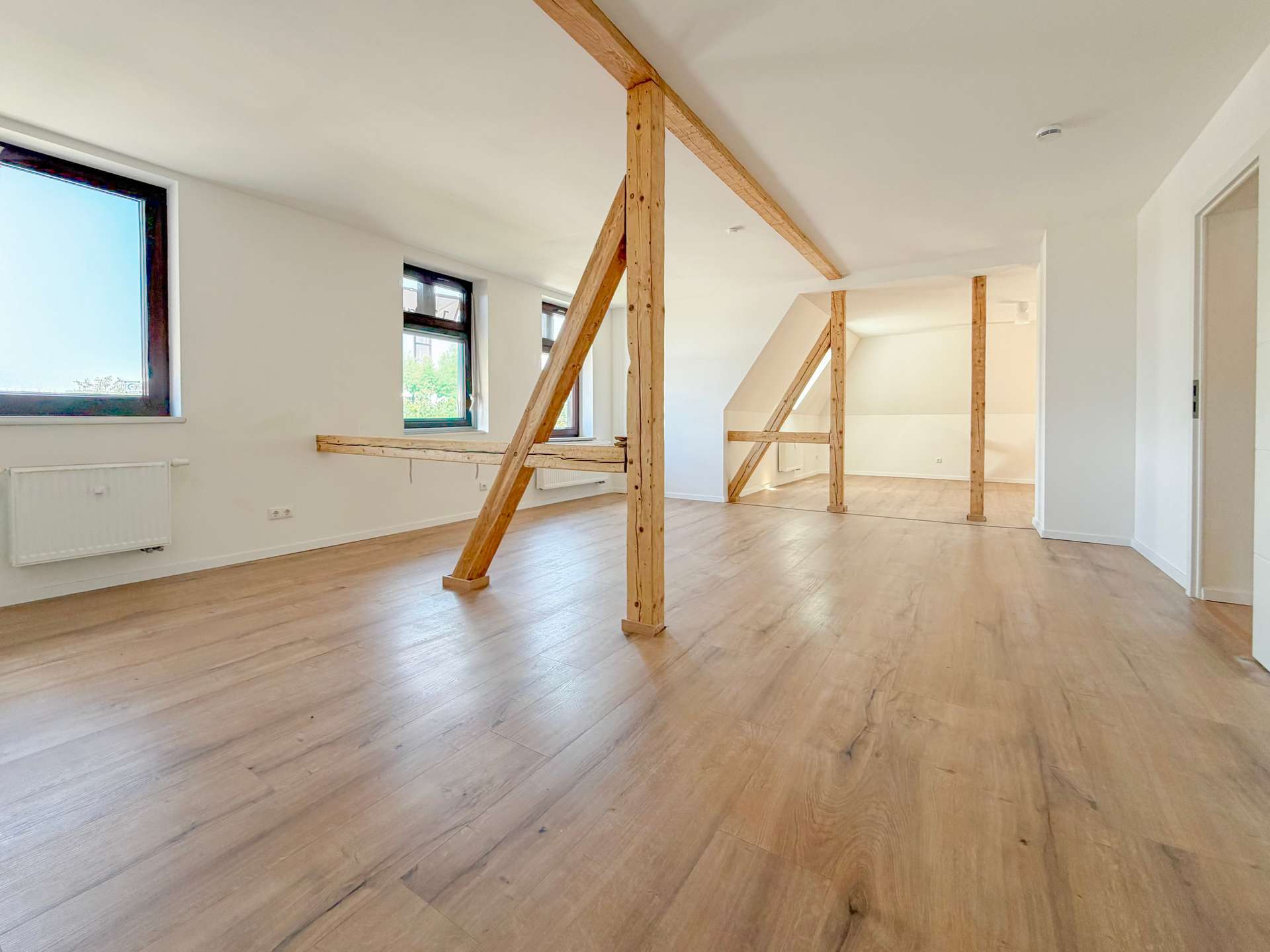Bright empty loft with light wood floors, white walls, and exposed wooden structural beams running across the ceiling and down the room edge.