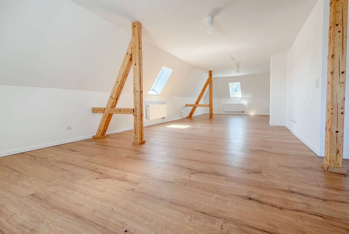Bright empty attic room with sloped white walls, exposed wooden beams, and light wood flooring; skylight windows bring natural light.