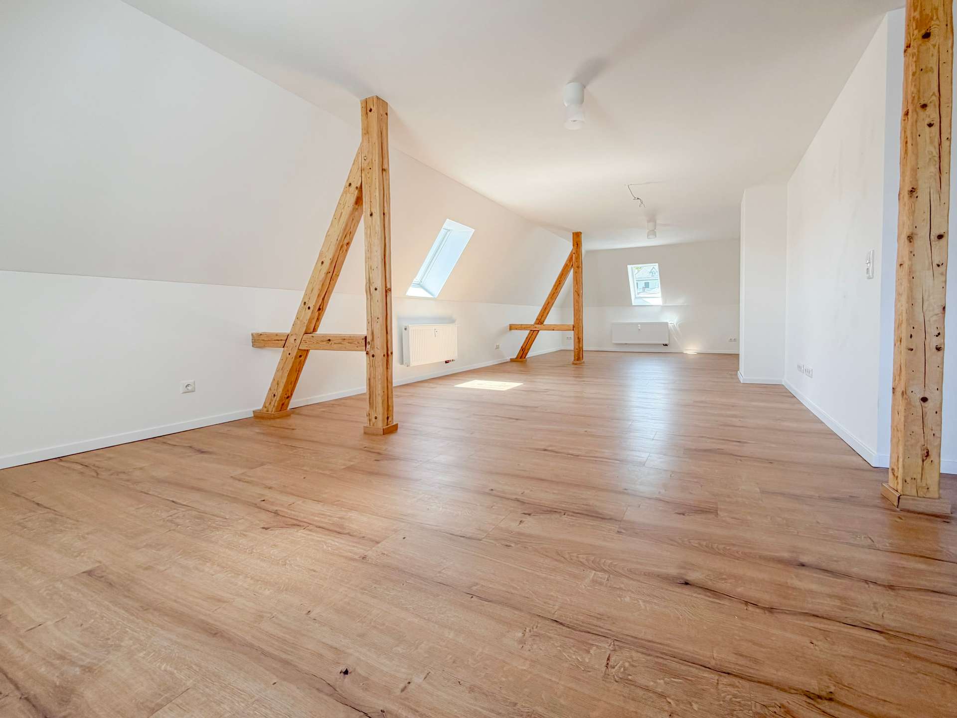 Bright empty attic room with sloped white walls, exposed wooden beams, and light wood flooring; skylight windows bring natural light.