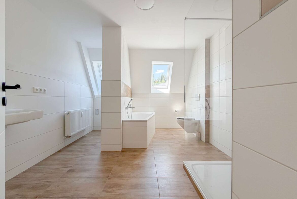 Bright modern bathroom with white tiles, freestanding bathtub, glass shower enclosure, and skylight window overlooking trees.