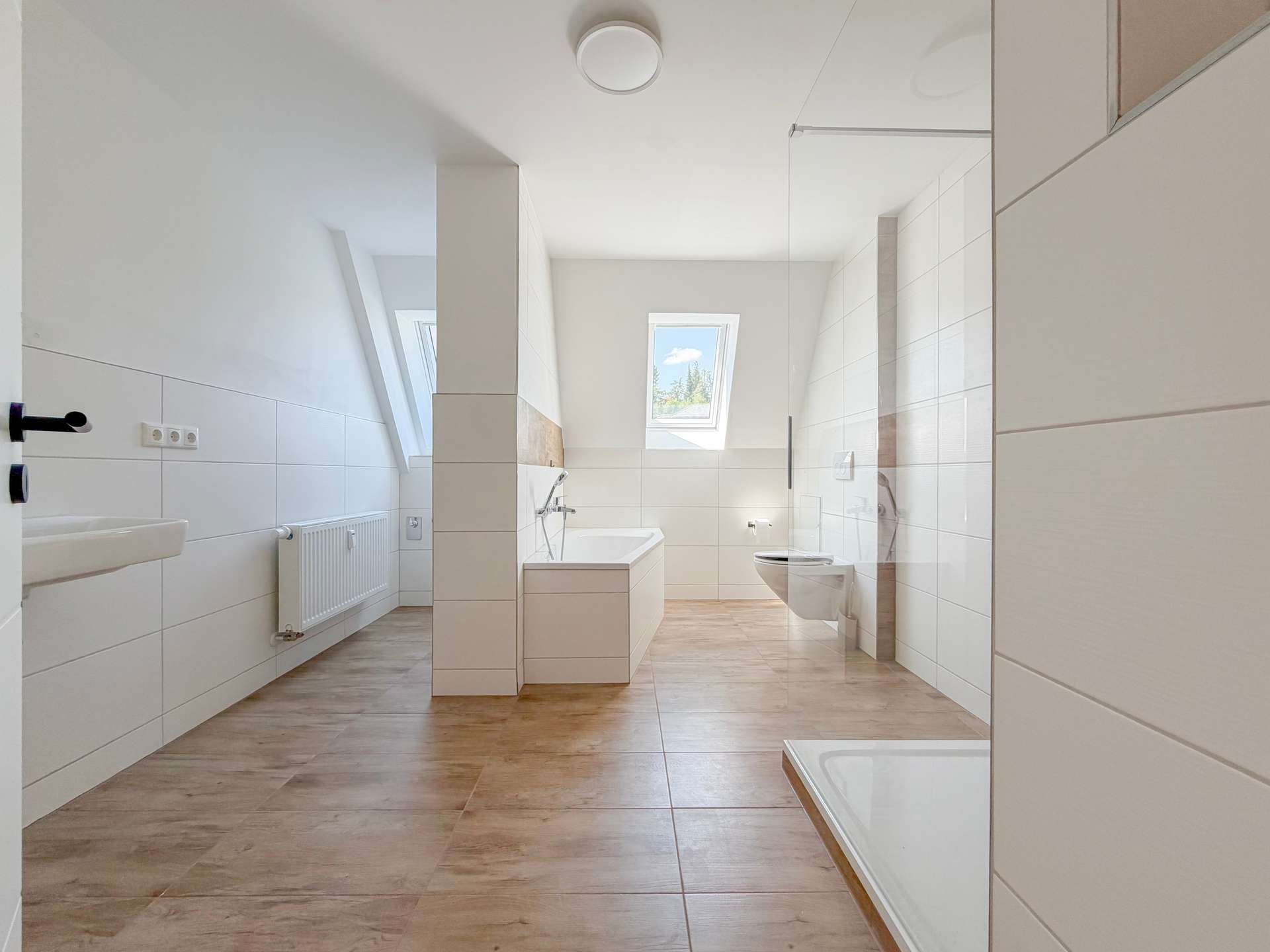 Bright modern bathroom with white tiles, freestanding bathtub, glass shower enclosure, and skylight window overlooking trees.