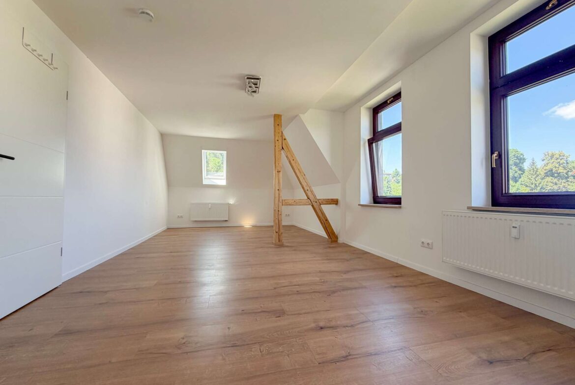 Bright, empty attic room with light wood flooring, sloped ceiling, and a vertical wooden beam near the center; large windows reveal greenery outside.