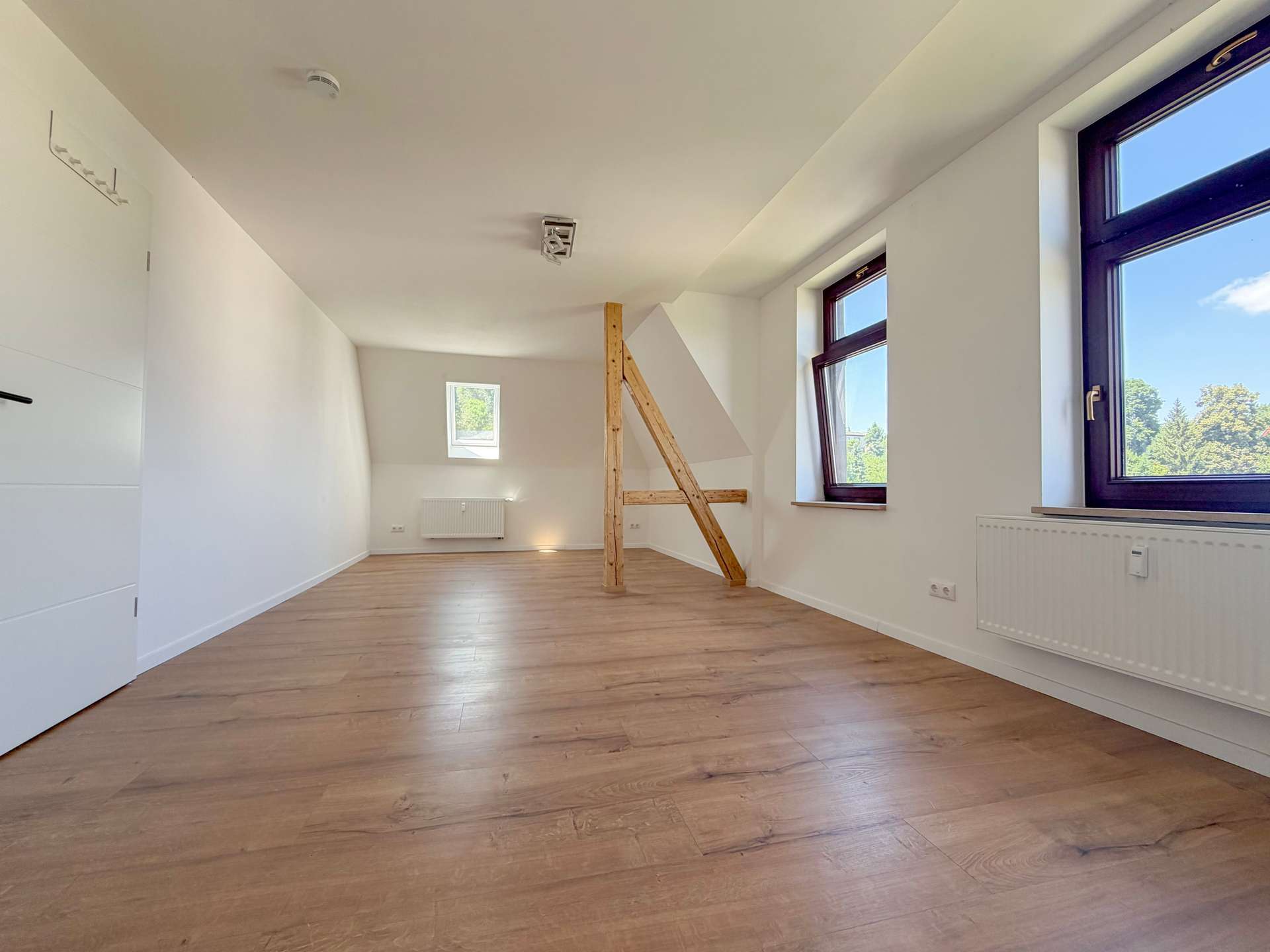 Bright, empty attic room with light wood flooring, sloped ceiling, and a vertical wooden beam near the center; large windows reveal greenery outside.
