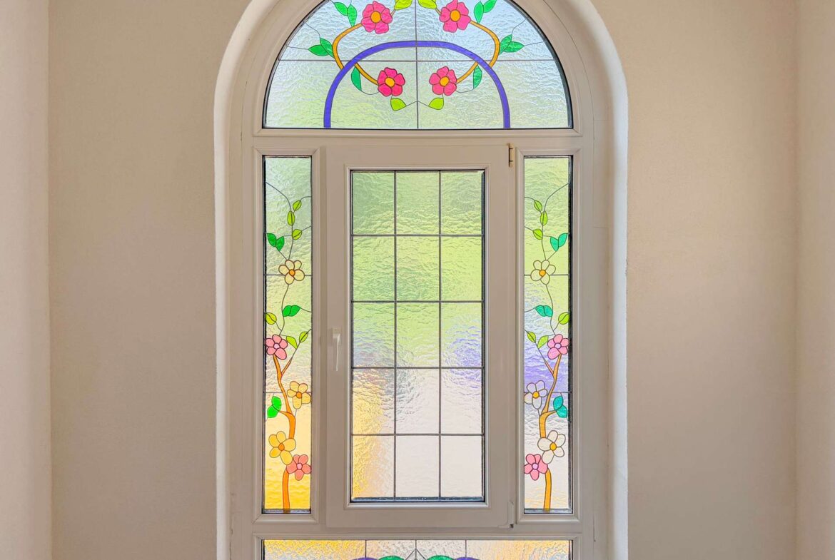 Arched decorative stained-glass window with pink flowers and green leaves set in a white frame on a beige wall.