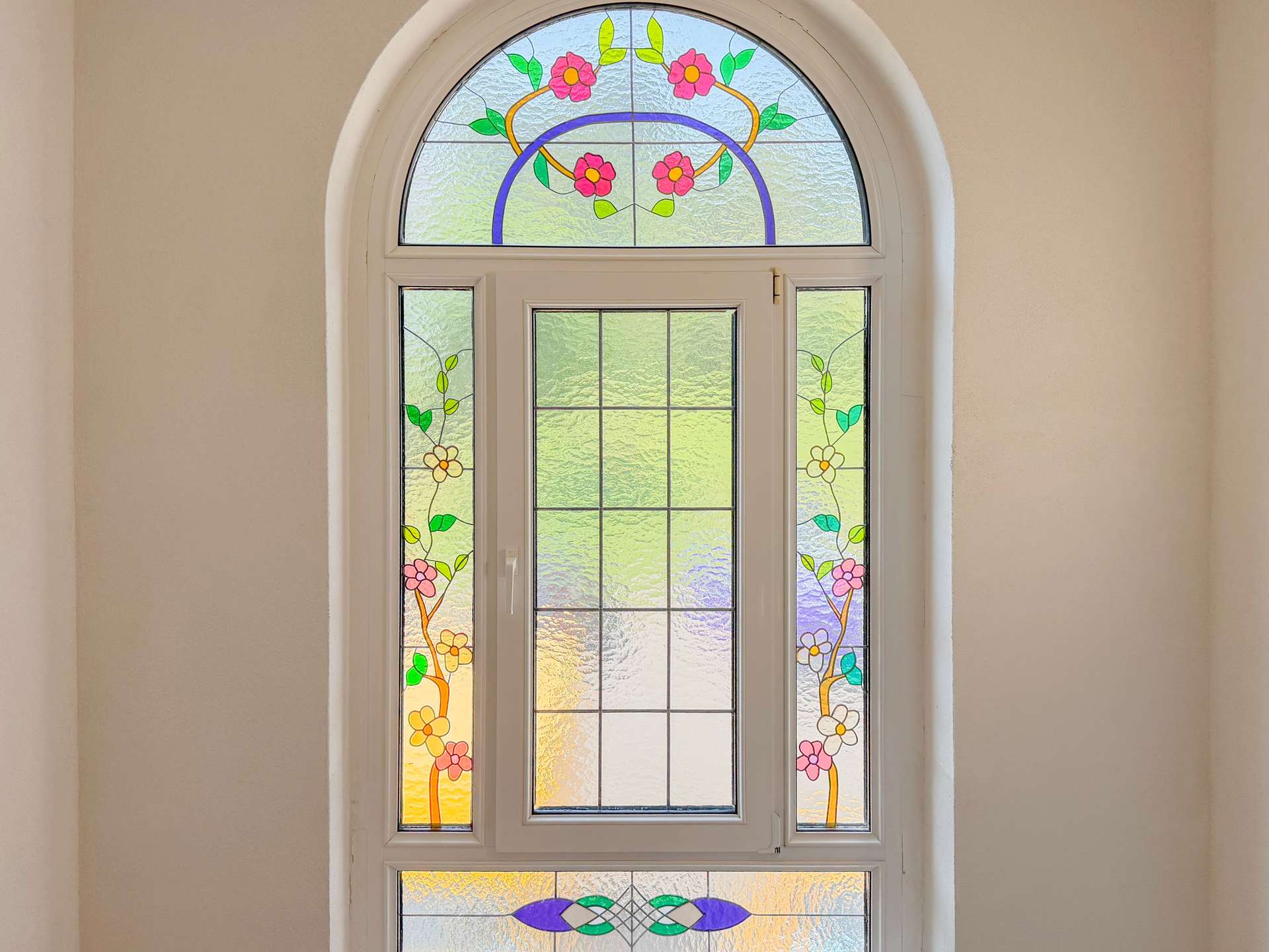 Arched decorative stained-glass window with pink flowers and green leaves set in a white frame on a beige wall.