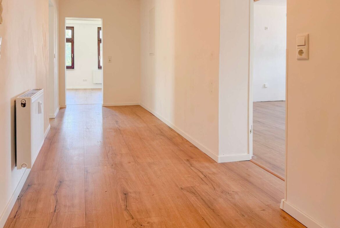 Bright hallway in a home with wood flooring, white walls, a radiator on the left, and open doorways showing a sunlit room at the end.