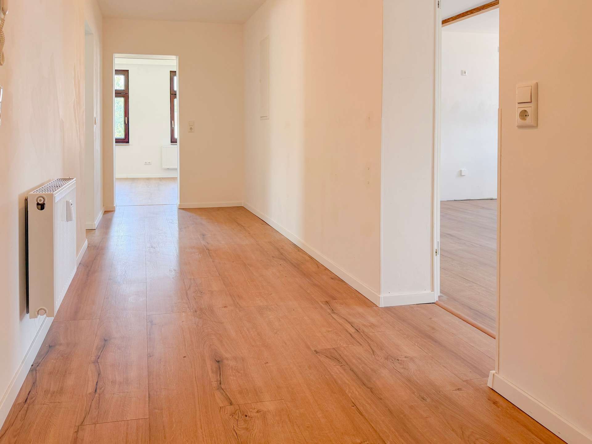 Bright hallway in a home with wood flooring, white walls, a radiator on the left, and open doorways showing a sunlit room at the end.