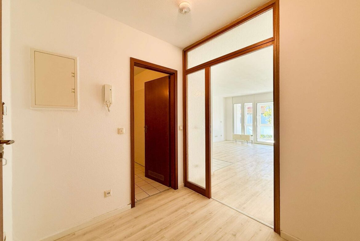 Hallway with light wood flooring and white walls, a wooden door and a glass-framed entry leading to a bright living area with large windows in the background.