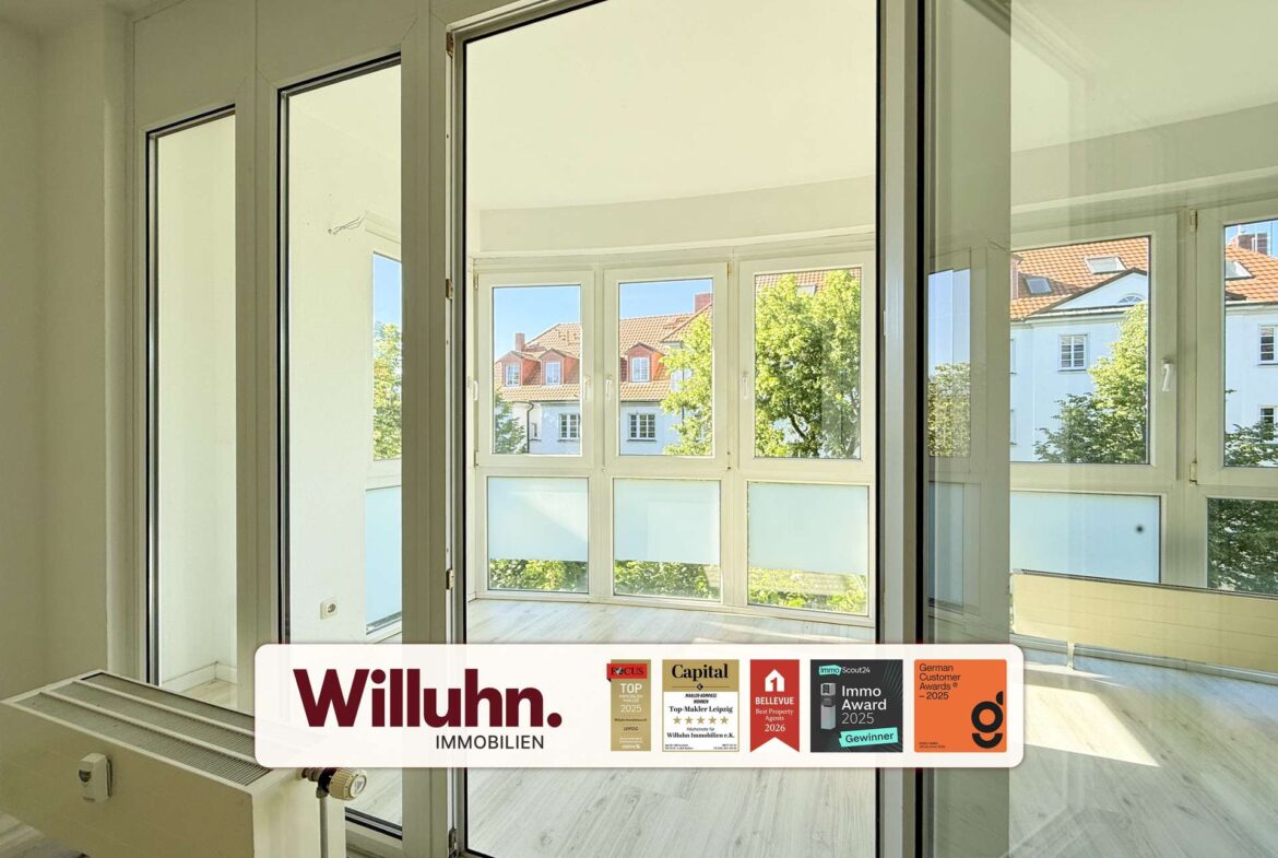 Interior of a bright room with large white-framed windows and a glass door opening to a balcony; exterior buildings visible.
