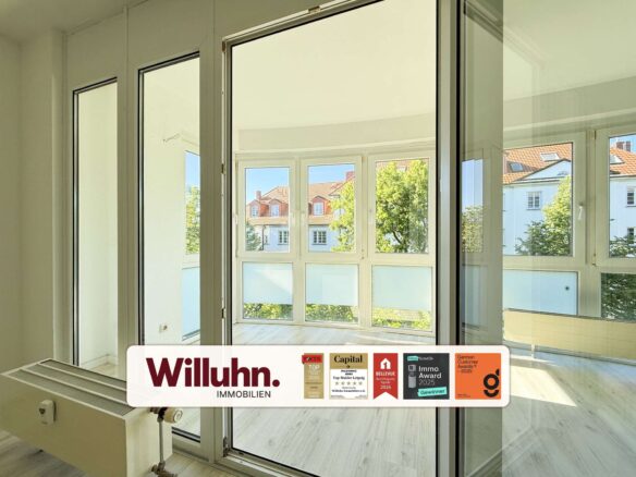 Interior of a bright room with large white-framed windows and a glass door opening to a balcony; exterior buildings visible.