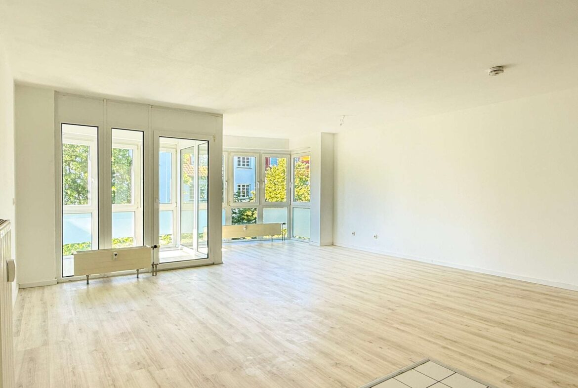 Bright, empty room with large glass doors and windows, light wood flooring, and white walls; radiator along the left wall and leafy outdoor view.
