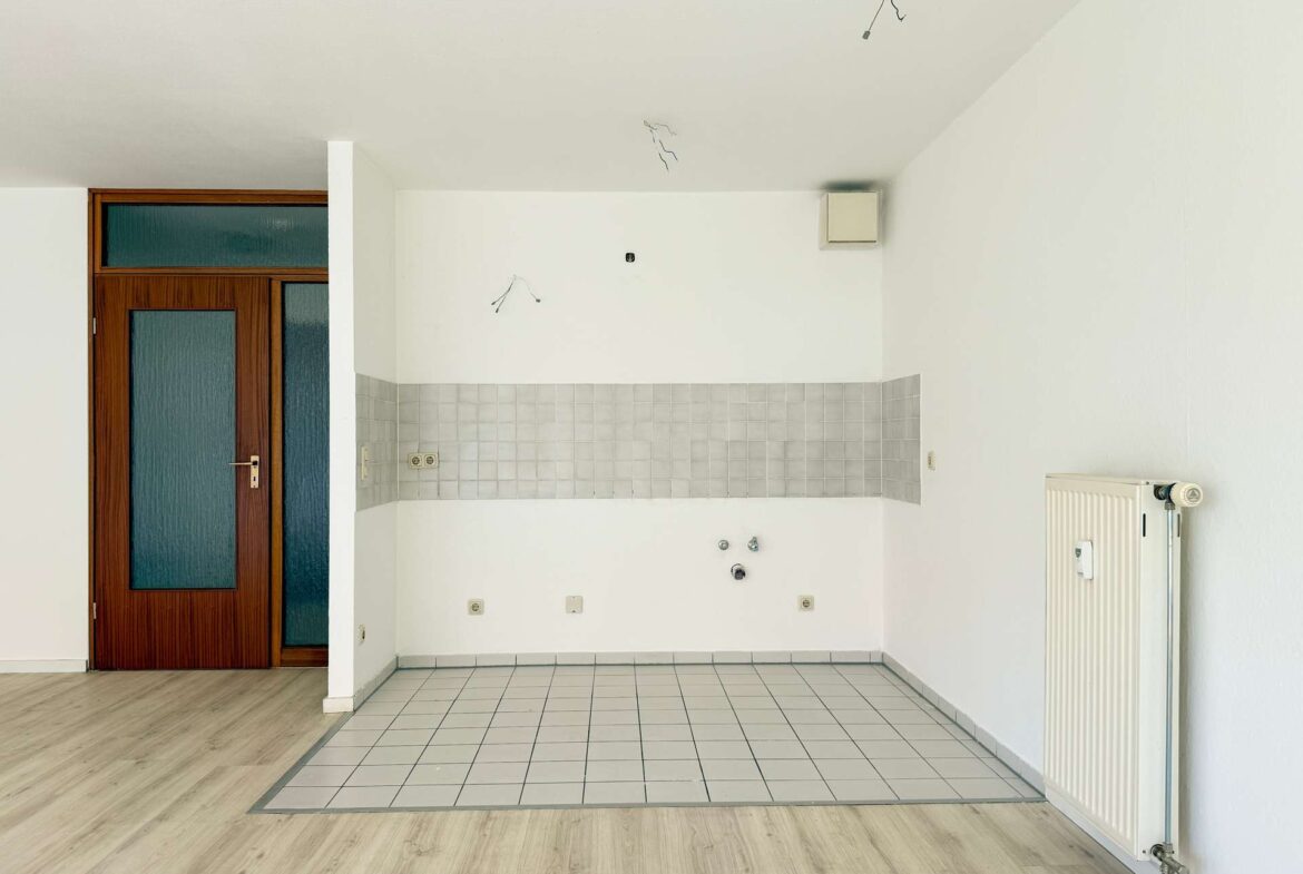 Unfinished kitchen area with white tiled backsplash, no cabinets, exposed ceiling wires, wooden door on left, radiator on right.