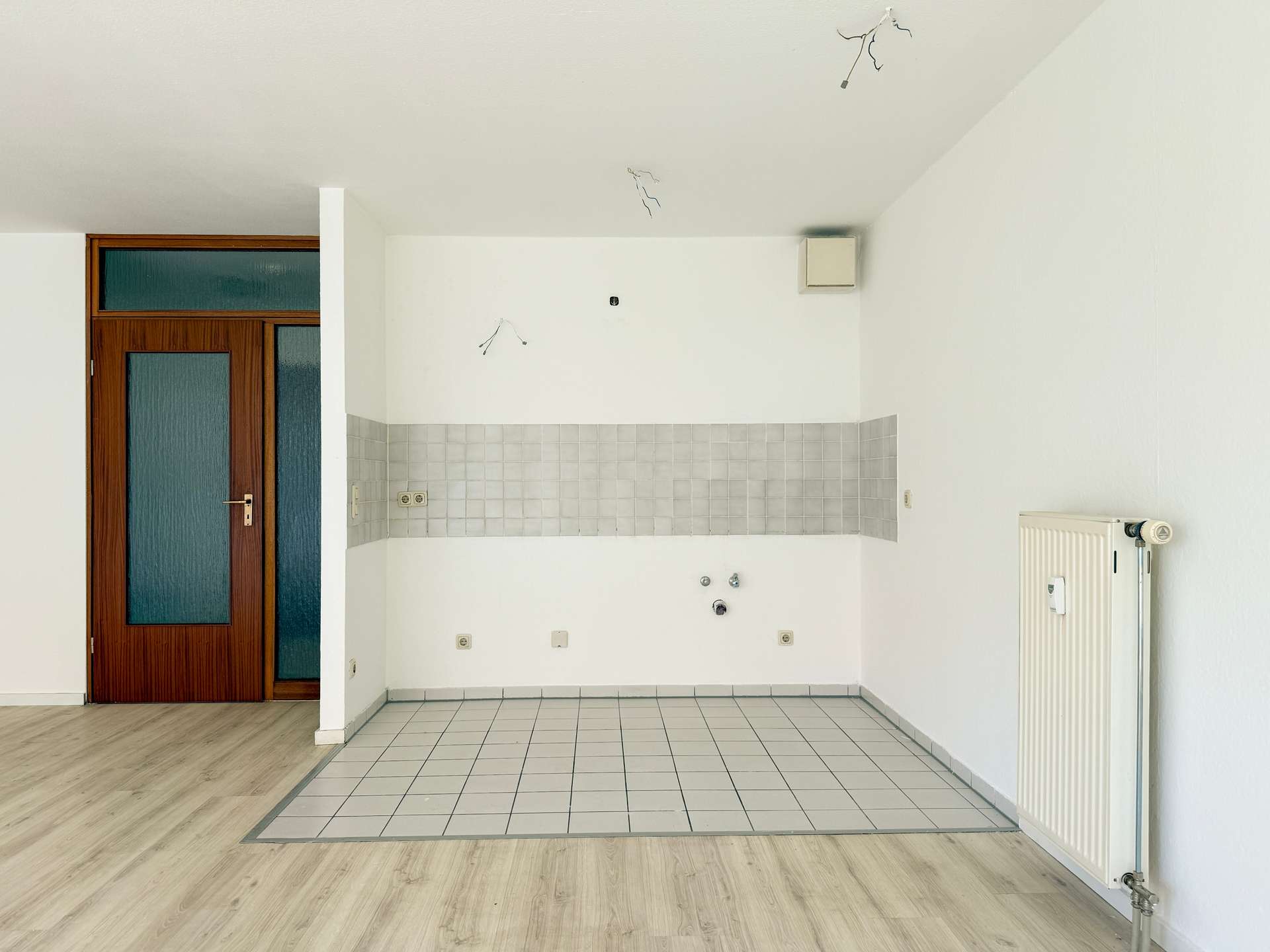 Unfinished kitchen area with white tiled backsplash, no cabinets, exposed ceiling wires, wooden door on left, radiator on right.