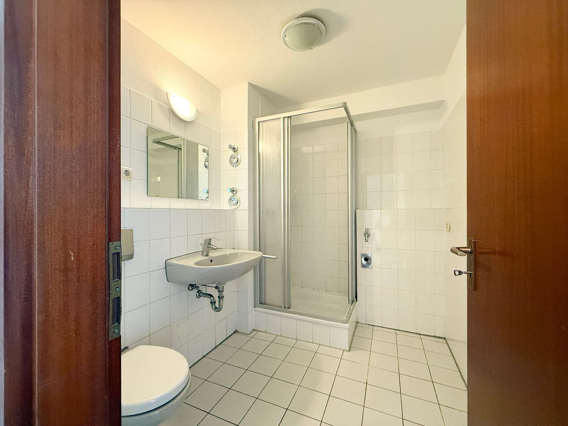 Small tiled bathroom featuring a wall-mounted sink, toilet, and a glass shower enclosure seen through an open door frame.
