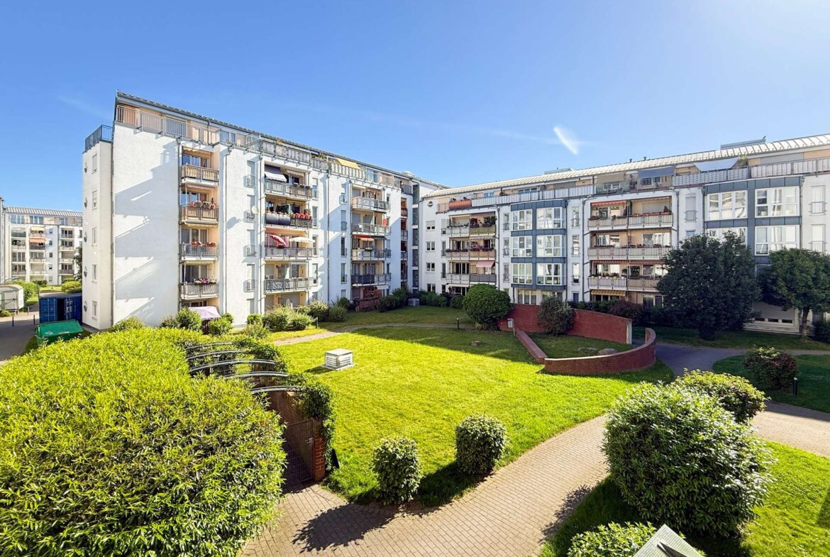 Residential apartment complex surrounding a green courtyard with hedges and paved paths under a blue sky.
