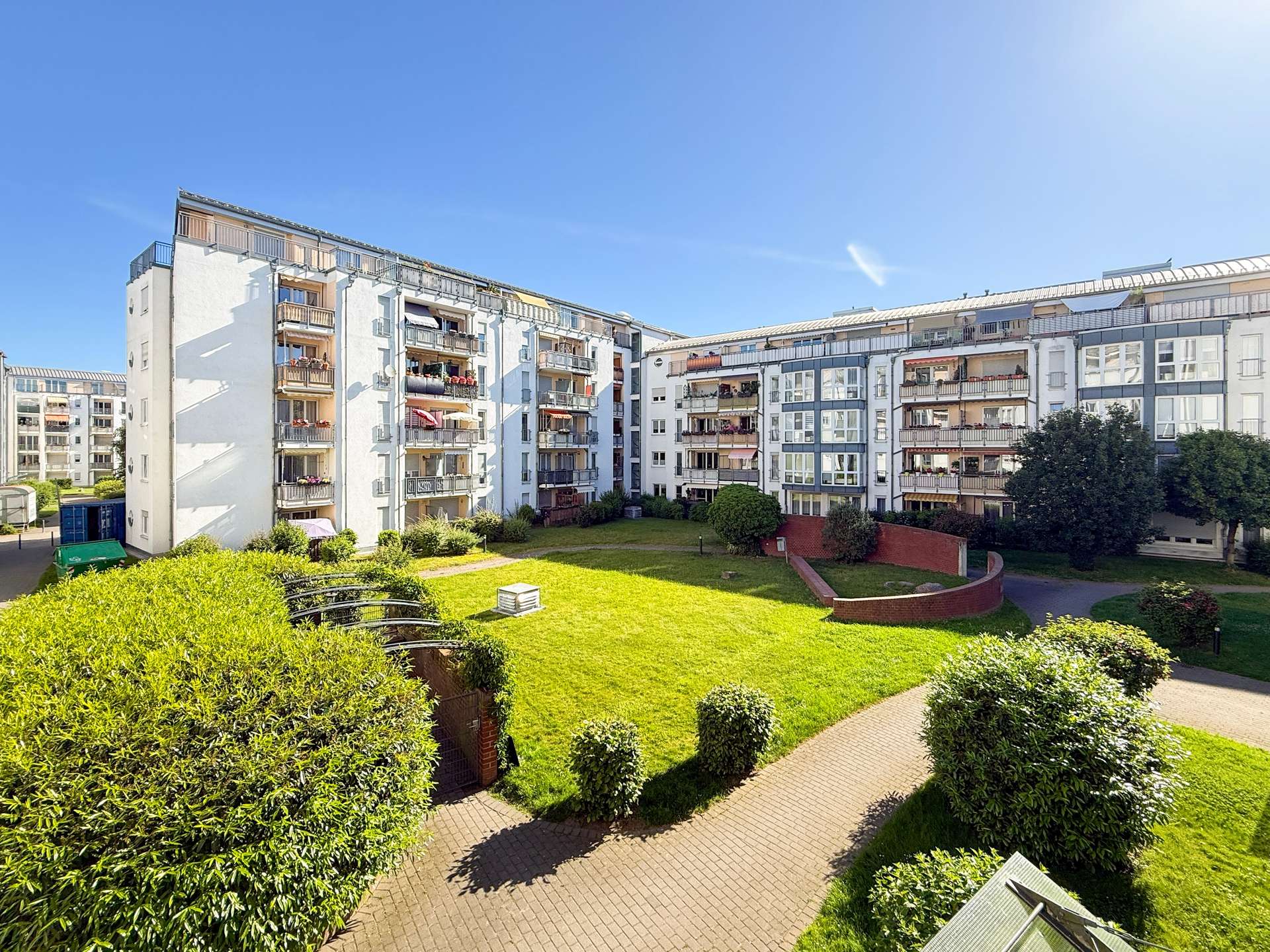 Residential apartment complex surrounding a green courtyard with hedges and paved paths under a blue sky.