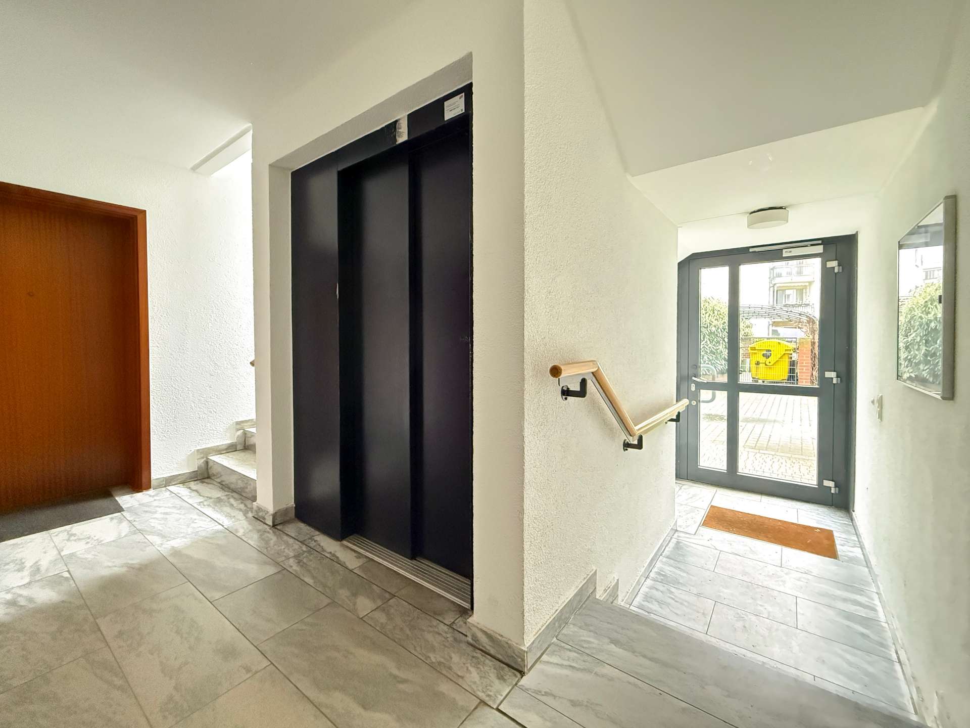 Interior hallway with a black elevator door alcove, a staircase with a wooden handrail on the right, and a glass exit door ahead.