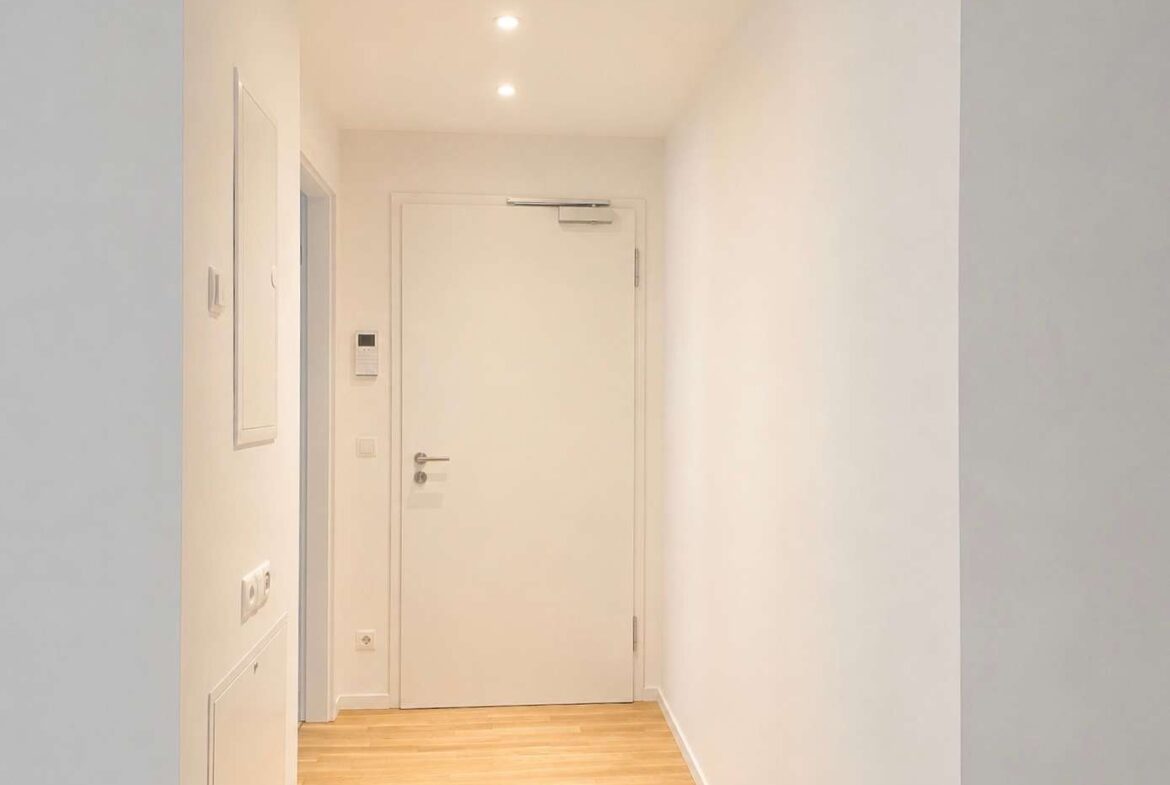 Bright white hallway with a closed white door at the end, warm wooden floor and recessed ceiling lights.