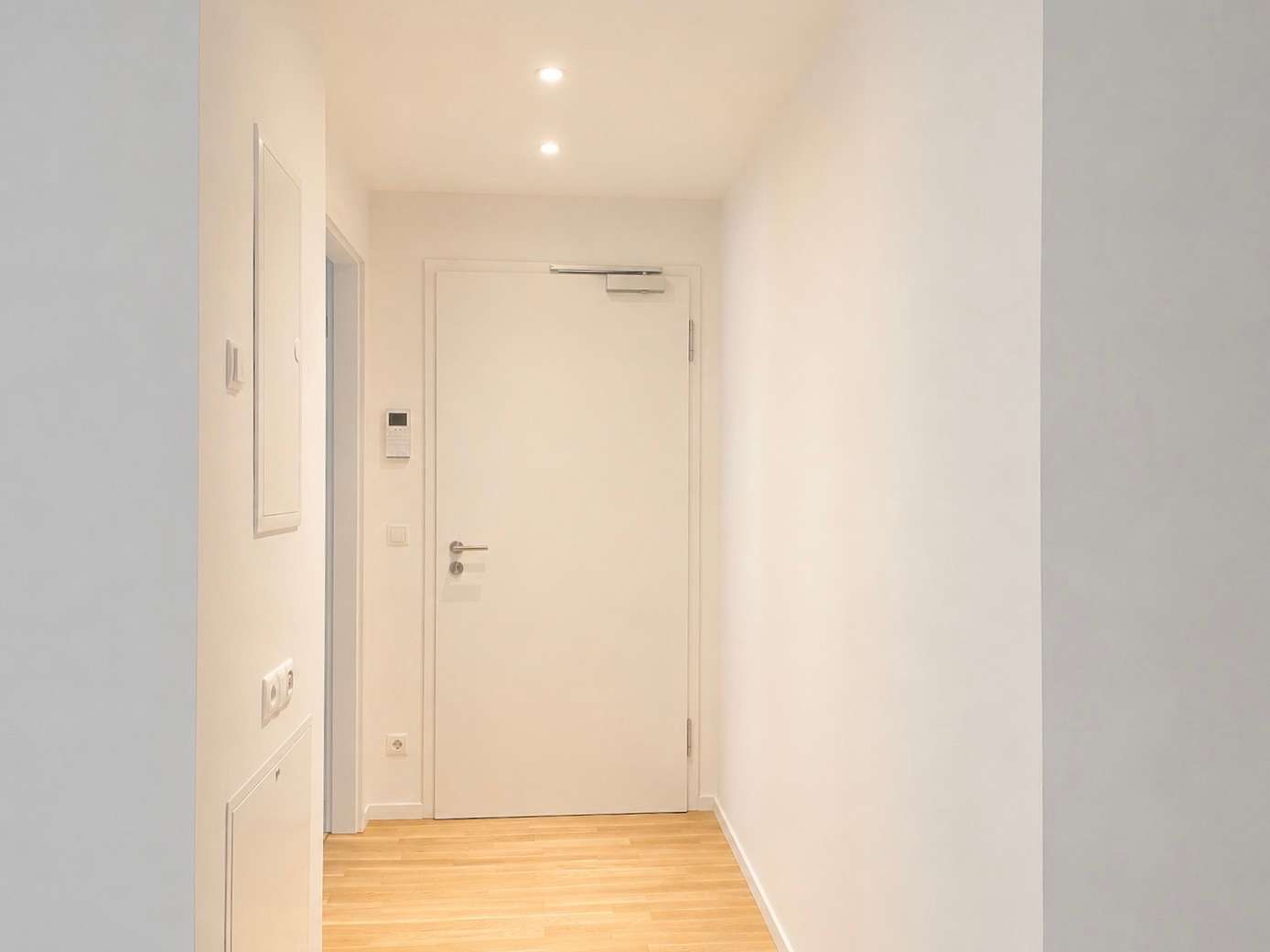 Bright white hallway with a closed white door at the end, warm wooden floor and recessed ceiling lights.