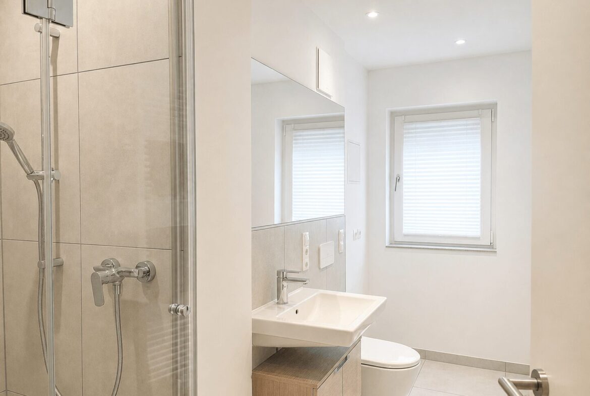 Modern bathroom with a glass shower enclosure on the left, chrome fixtures, a floating sink, and a window with white blinds. A toilet sits beside the sink under a large mirror.