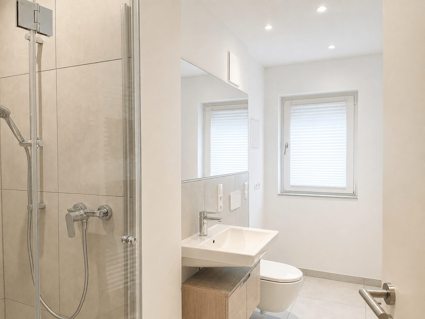 Modern bathroom with a glass shower enclosure on the left, chrome fixtures, a floating sink, and a window with white blinds. A toilet sits beside the sink under a large mirror.