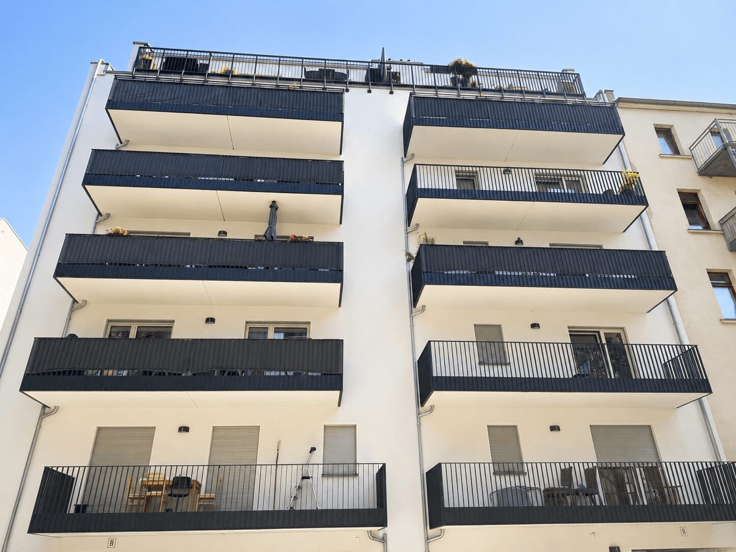 White apartment building with multiple blue-railed balconies stacked on two towers against a clear blue sky.