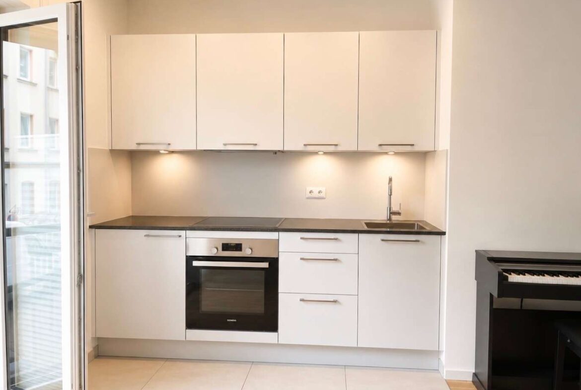 Modern white kitchen with upper and lower cabinets, built-in oven, stovetop, sink, and under-cabinet lighting.