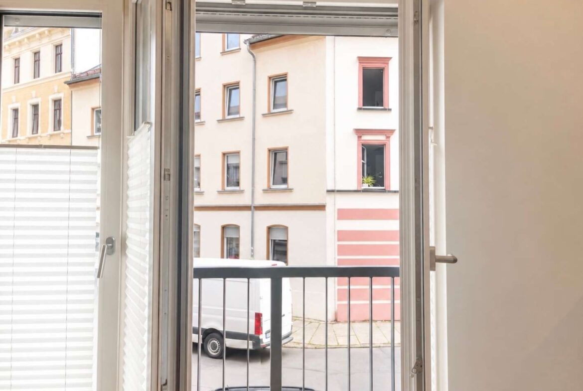 Open French doors with white blinds frame a view of a balcony overlooking a street with a parked white van and pastel-colored buildings across the road.