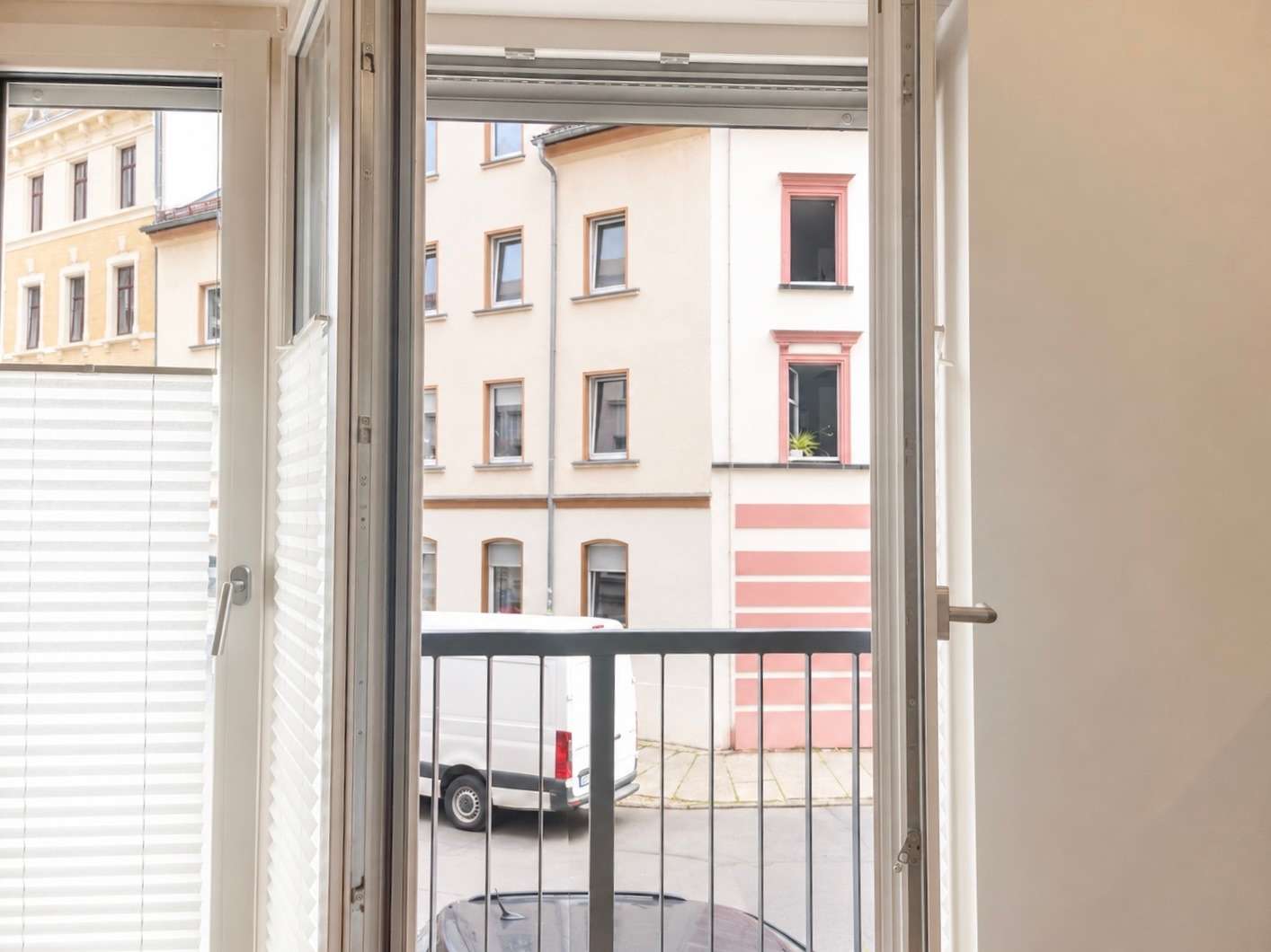 Open French doors with white blinds frame a view of a balcony overlooking a street with a parked white van and pastel-colored buildings across the road.
