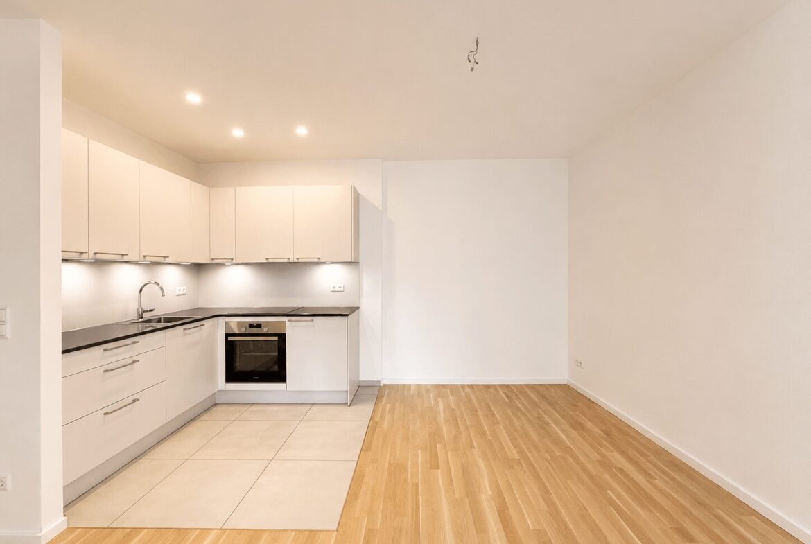 Open-plan kitchen with white cabinets, built-in oven, and gray countertops along a tiled section transitioning to wood flooring in a bright, empty room
