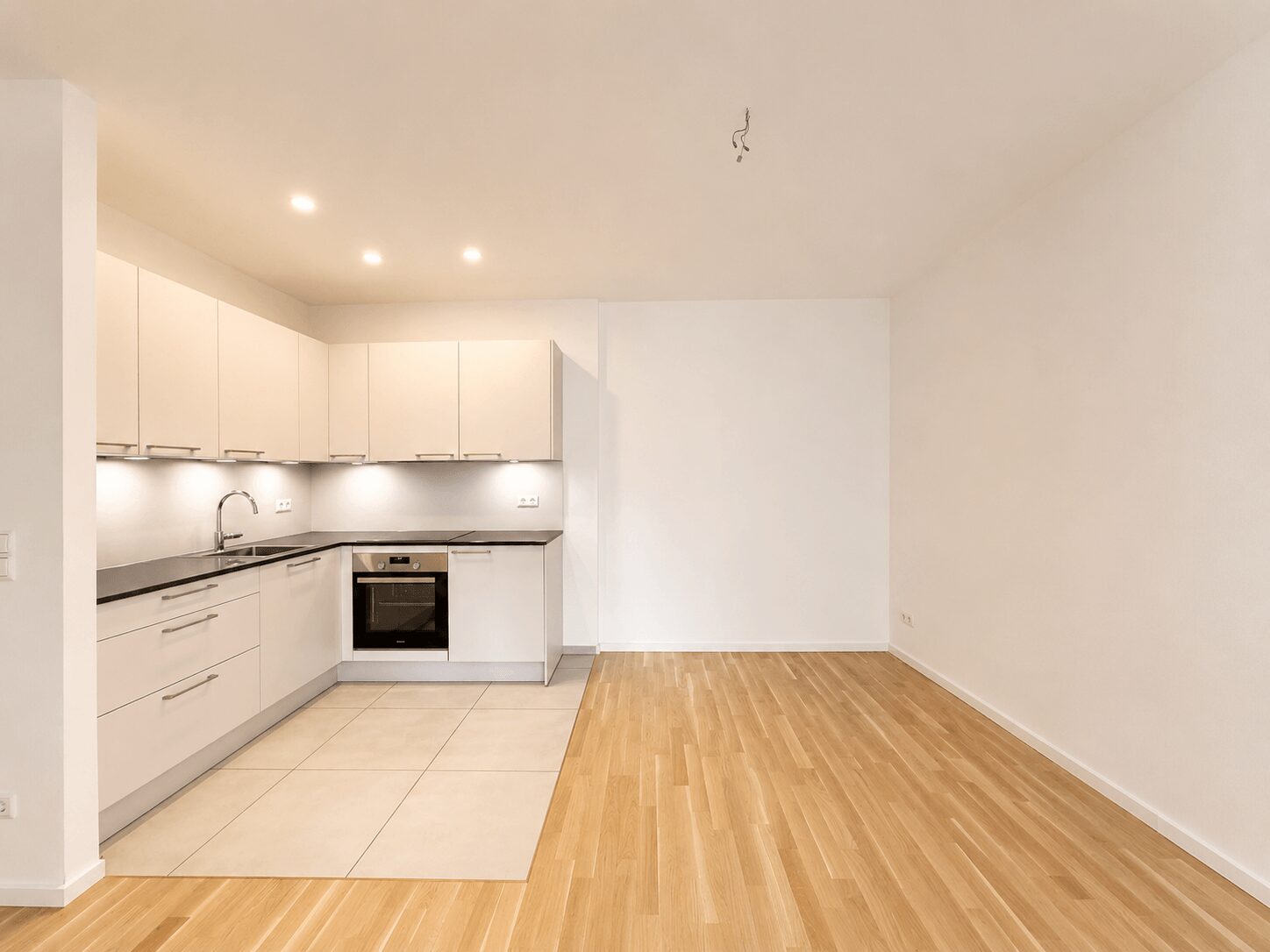 Open-plan kitchen with white cabinets, built-in oven, and gray countertops along a tiled section transitioning to wood flooring in a bright, empty room