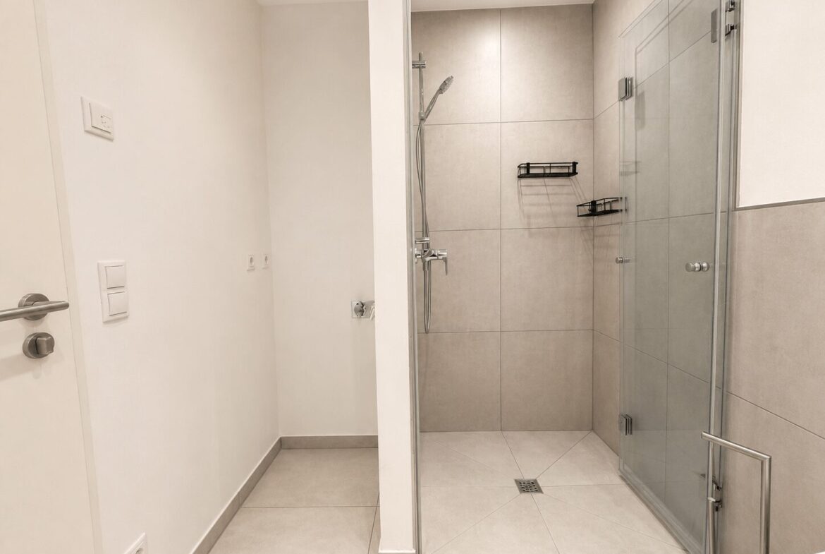 Modern bathroom entrance showing a glass shower enclosure with tiled walls and chrome fixtures.