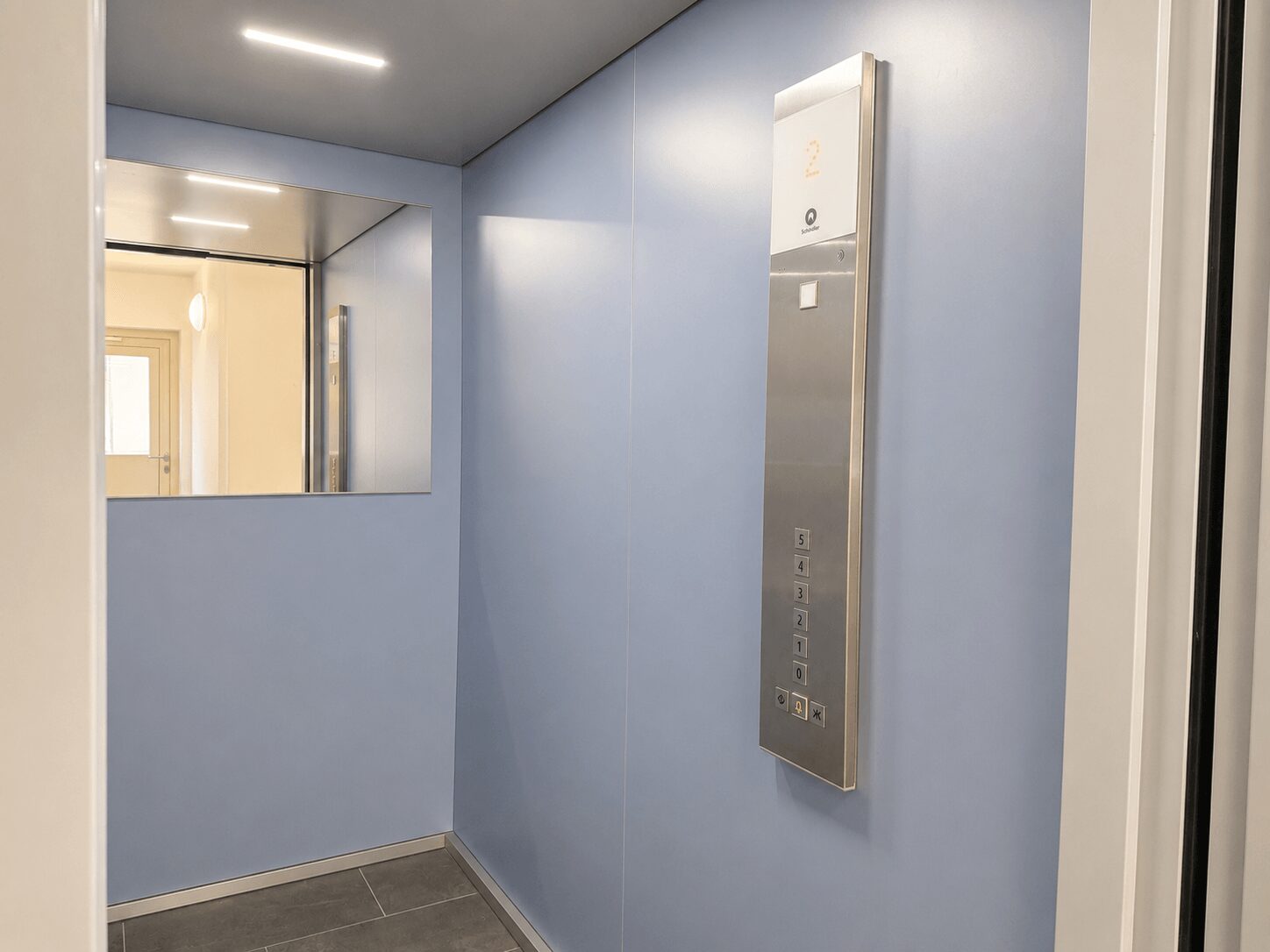 Elevator interior with light blue walls, a mirrored wall, and a stainless vertical control panel showing floor buttons 0–5.