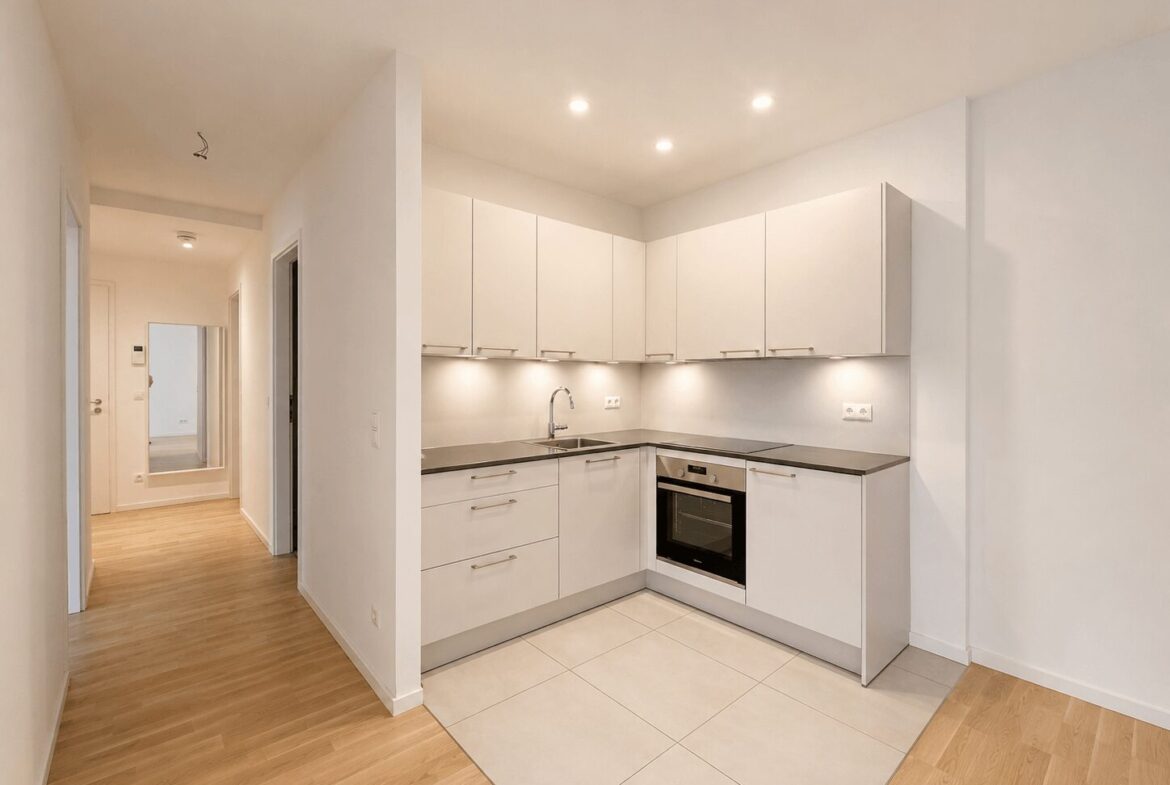 Modern white L-shaped kitchen with gray countertop, built-in oven, sink, and upper cabinets; hallway with mirror in background.