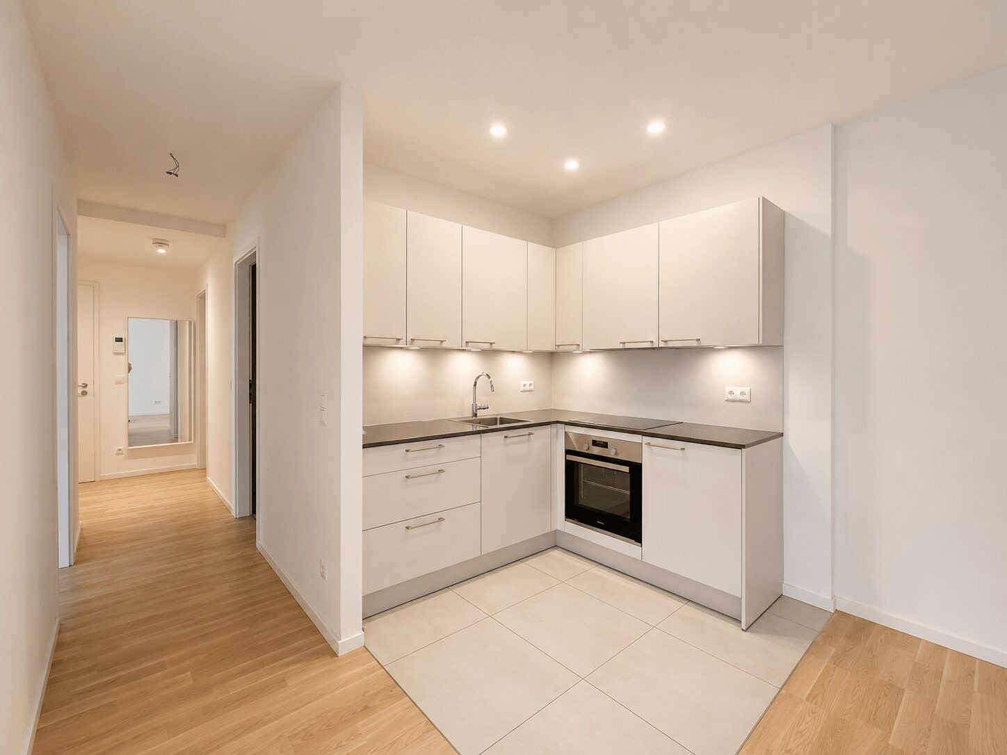 Modern white L-shaped kitchen with gray countertop, built-in oven, sink, and upper cabinets; hallway with mirror in background.