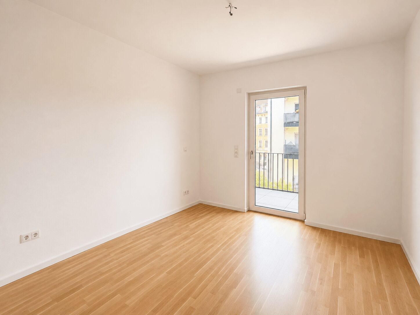 Empty white-walled room with light wood floor and a glass door opening to a small balcony with a metal railing outside.