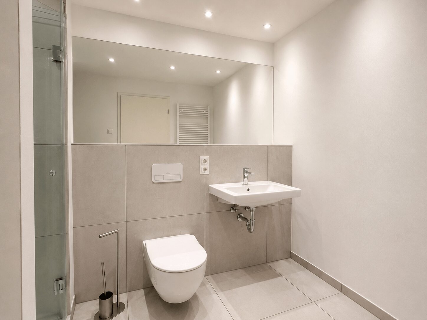 Modern bathroom with wall-mounted toilet, rectangular sink, large mirror, and glass-enclosed shower on the left.